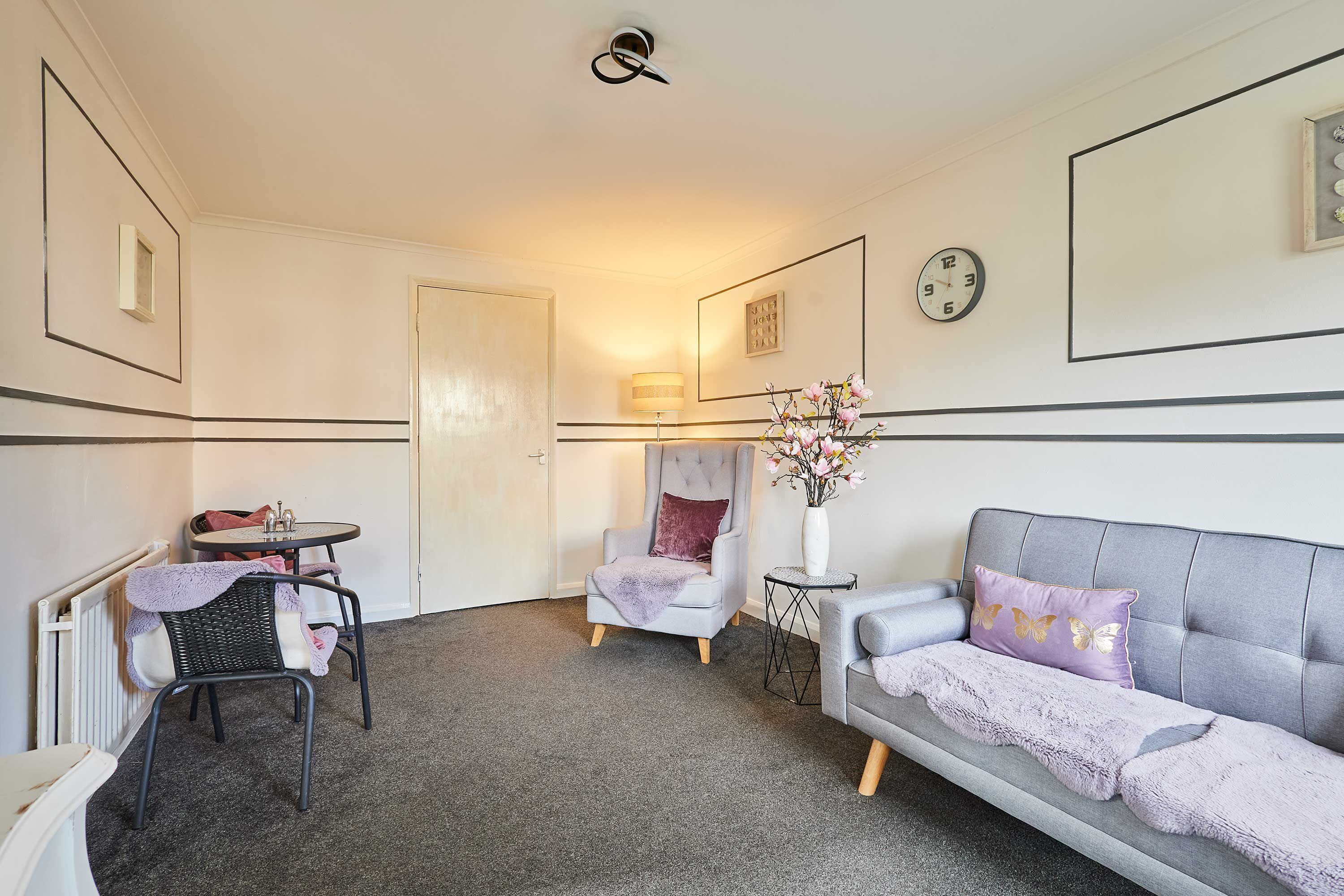 Une pièce comprenant un canapé gris, un fauteuil, une petite table, un radiateur et une porte.