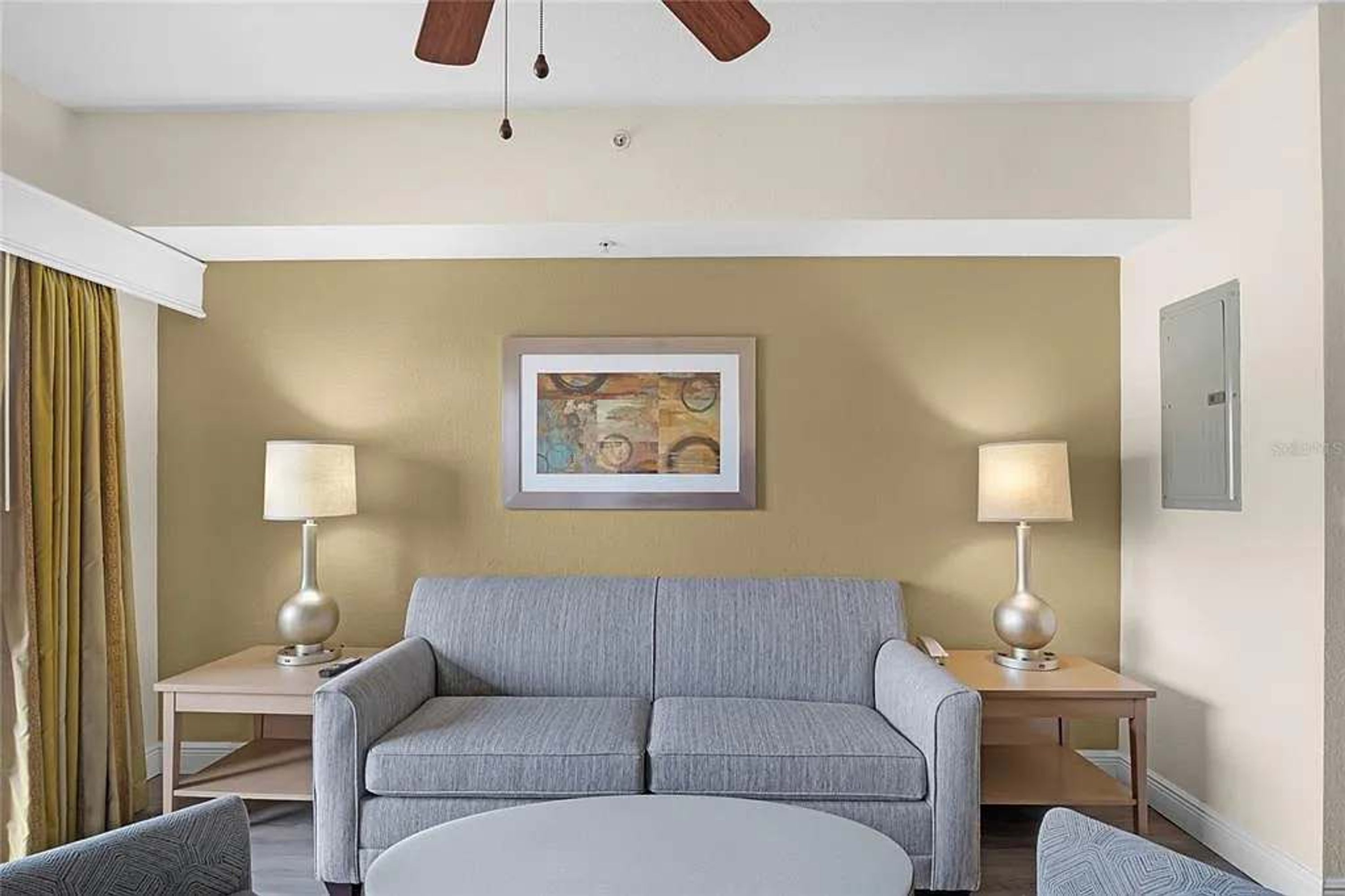 Quarto de hotel com sofá cinza, duas mesas de cabeceira com abajures, ventoinha no teto e uma obra de arte emoldurada na parede.