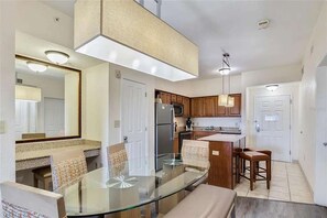 A modern kitchen with a glass dining table, wooden cabinets, and a built-in island. - Stay Near Disney 2BR Pools Arcade (Orlando)