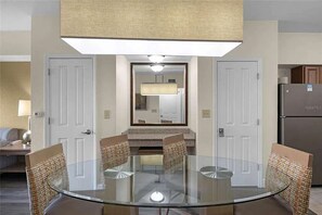 A dining area with a glass table surrounded by chairs, a kitchen with a refrigerator, and a mirror above the table. - Stay Near Disney 2BR Pools Arcade (Orlando)