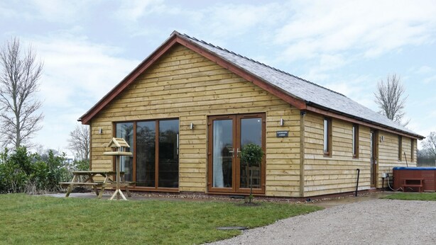 A wooden building with a pitched roof, large sliding glass doors, and a gravel driveway. - Gardener's Lodge (Nantwich)