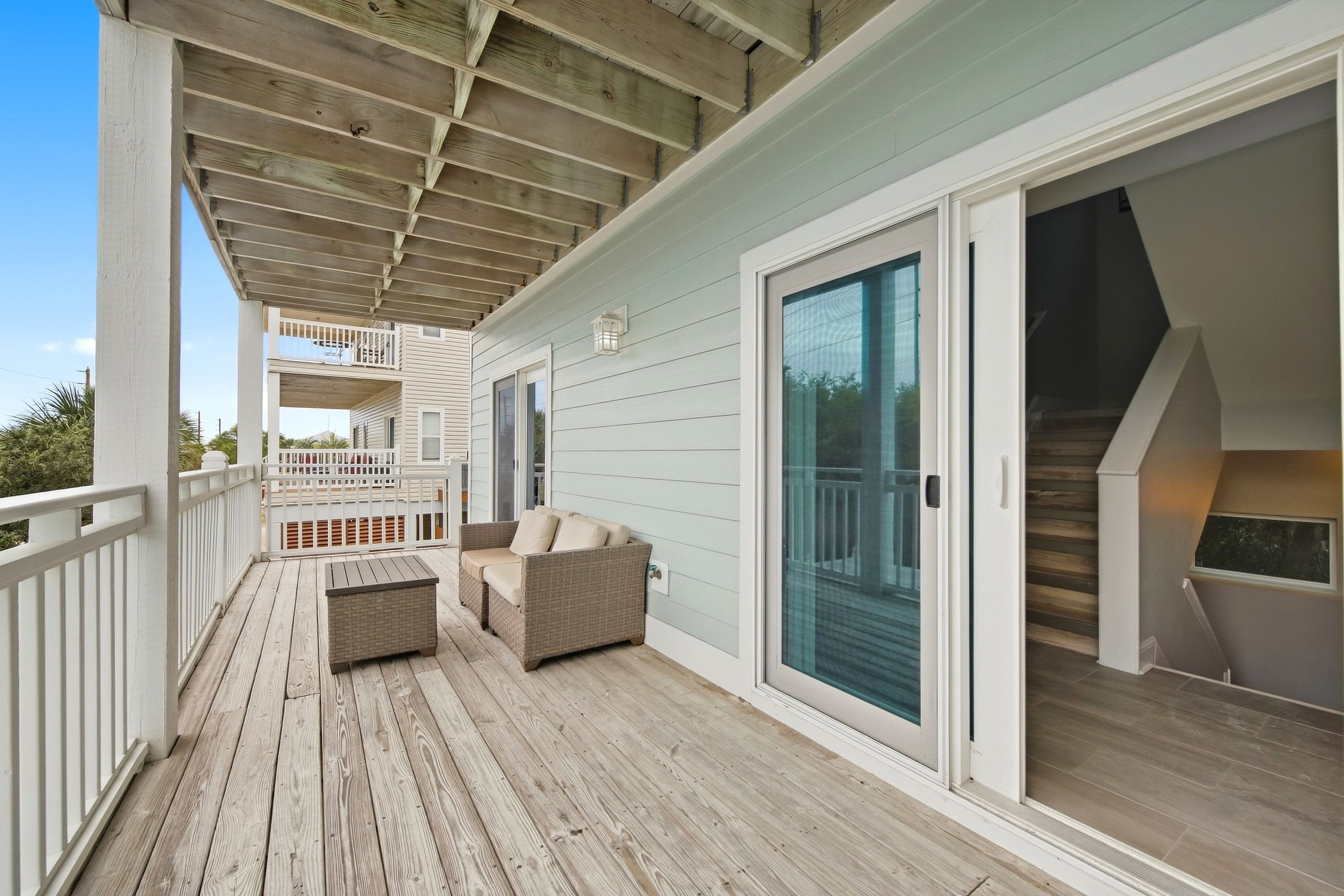 A covered outdoor deck with a sofa and a table, a sliding glass door, and a staircase leading to an upper level.