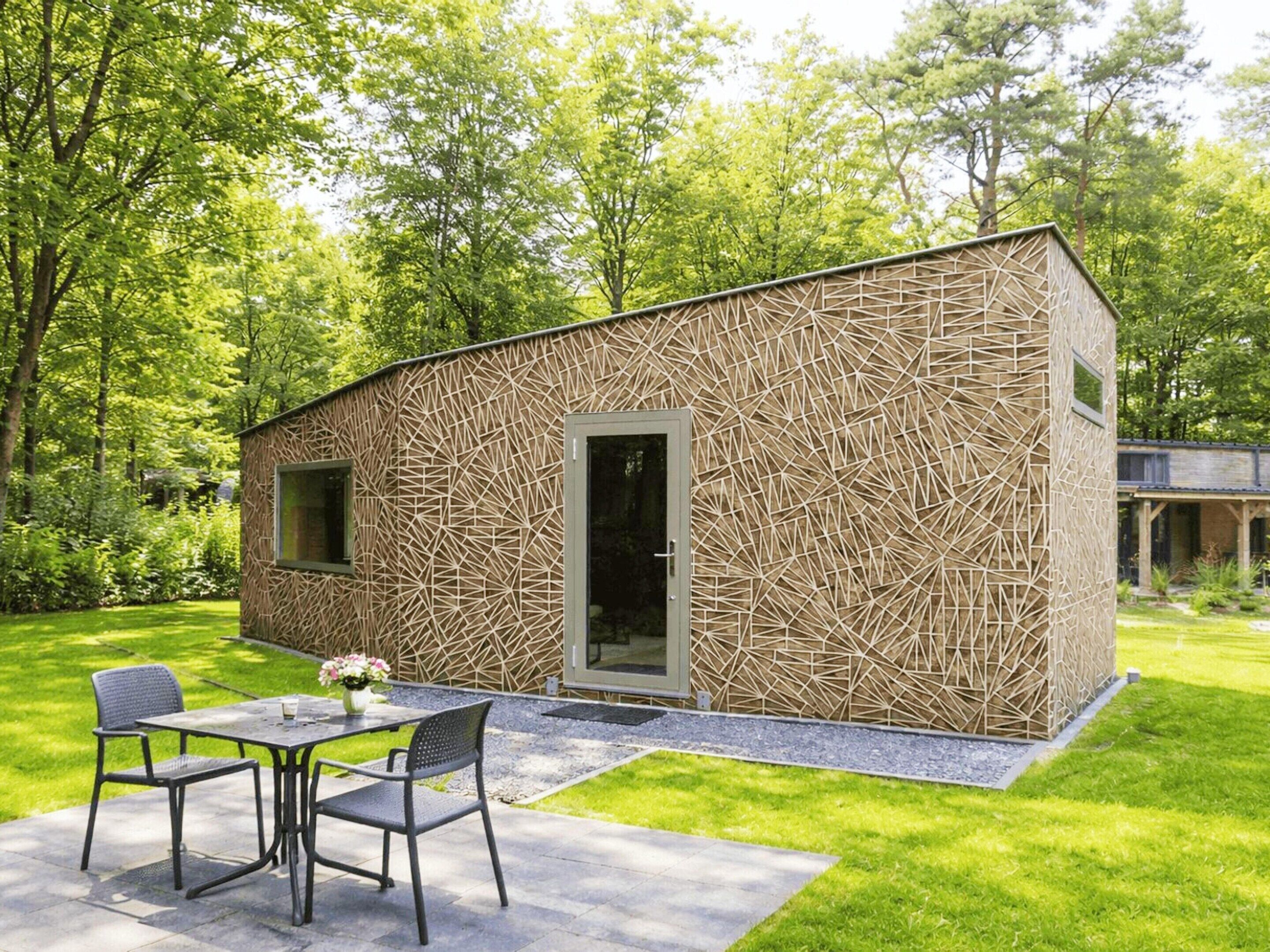 A modern outdoor seating area with a table and chairs, a small building with a geometric facade, and a lush green garden.