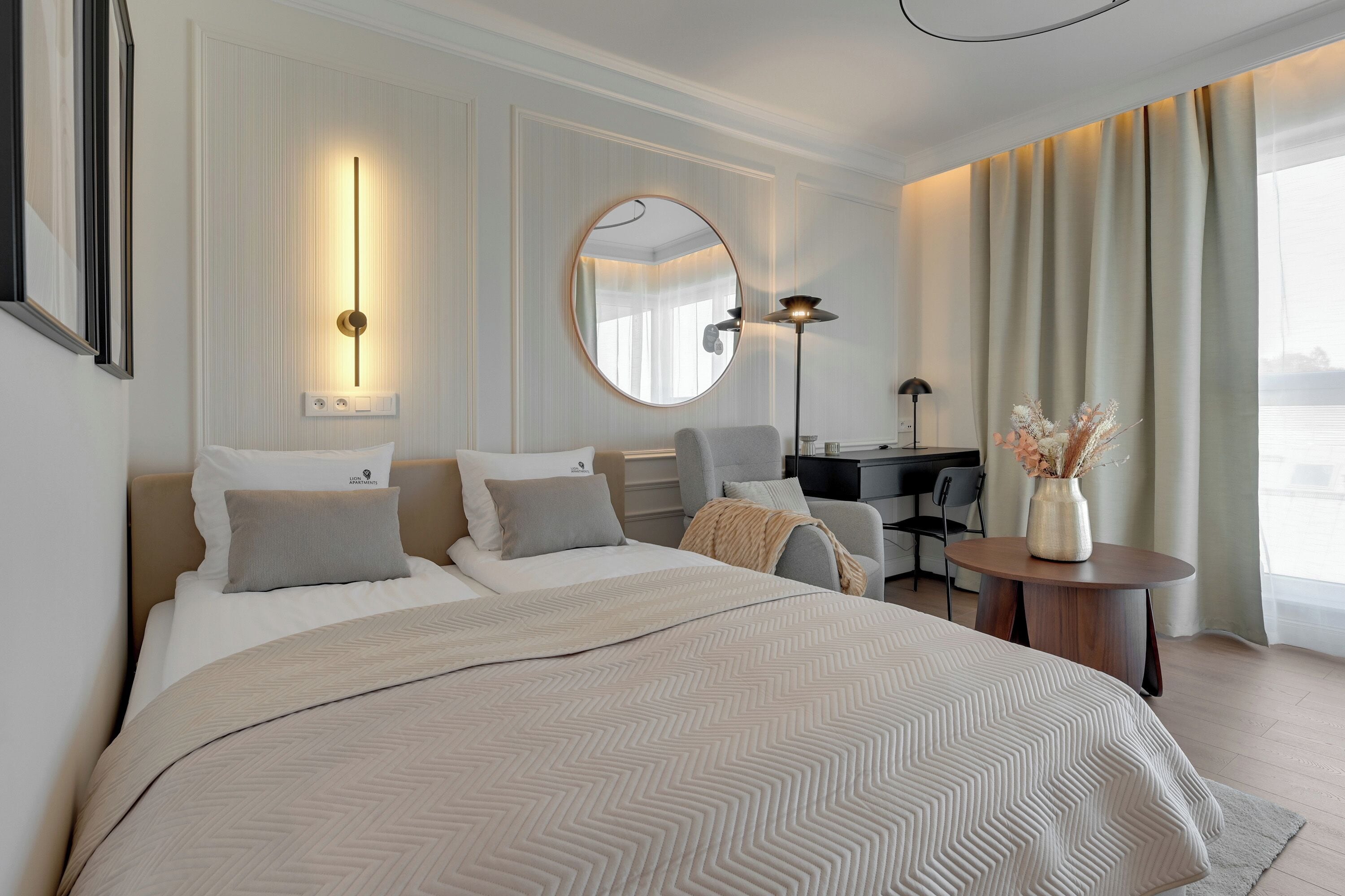 A modern hotel room with a large bed, a round mirror, a desk, and a view of the city.