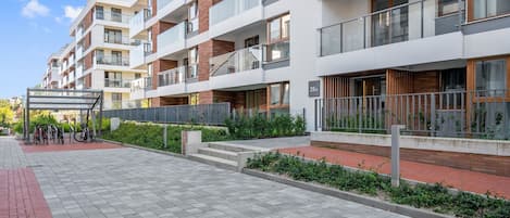 Un immeuble résidentiel à plusieurs étages, doté de balcons, d’un espace de stationnement pour vélos couvert et d’un chemin piétonnier pavé.