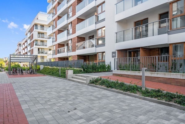 Un edificio residencial de varios pisos con balcones, una zona cubierta para aparcar bicicletas y un camino pavimentado.