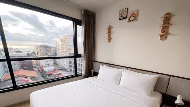 Habitación de hotel con una cama grande, una mesita de noche con lámpara y vista a la ciudad a través de un ventanal.