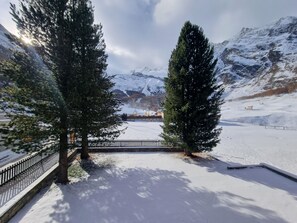 A snow-covered landscape with two prominent evergreen trees, a paved pathway, and a distant view of a building with a snow-covered roof. - Haus Rio Appartement/Fewo Appartement/Fewo by Interhome (Saas-Fee)