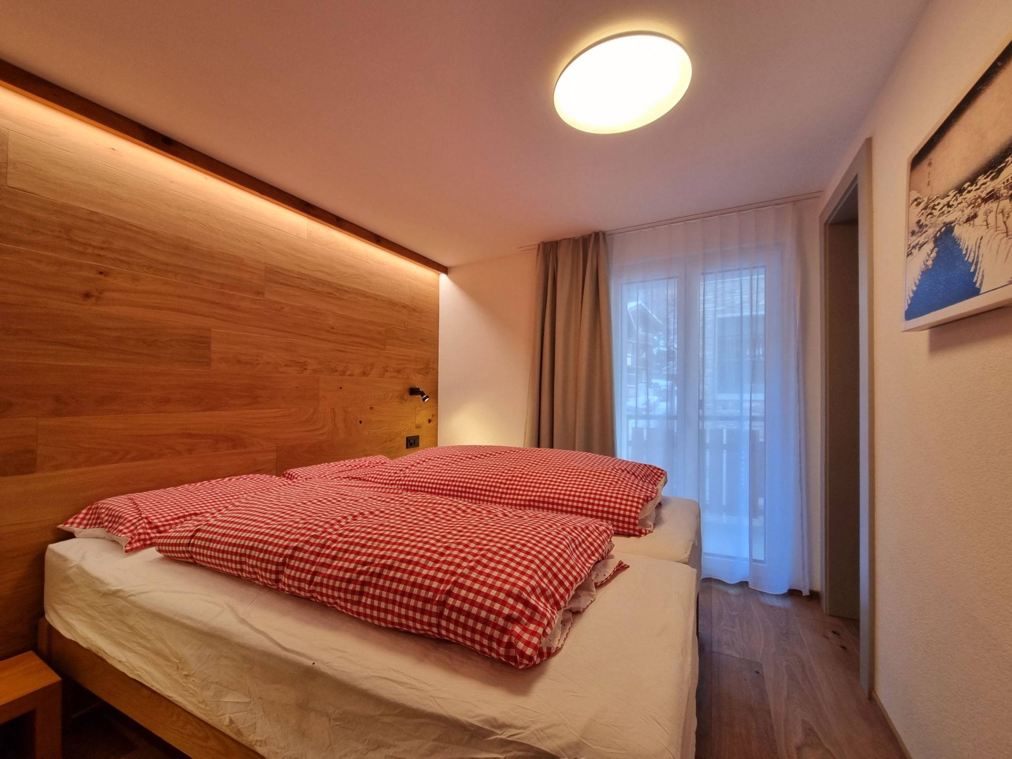 A bedroom with a wooden headboard, a bed with red and white checkered bedding, a bedside lamp, and a window with sheer curtains.