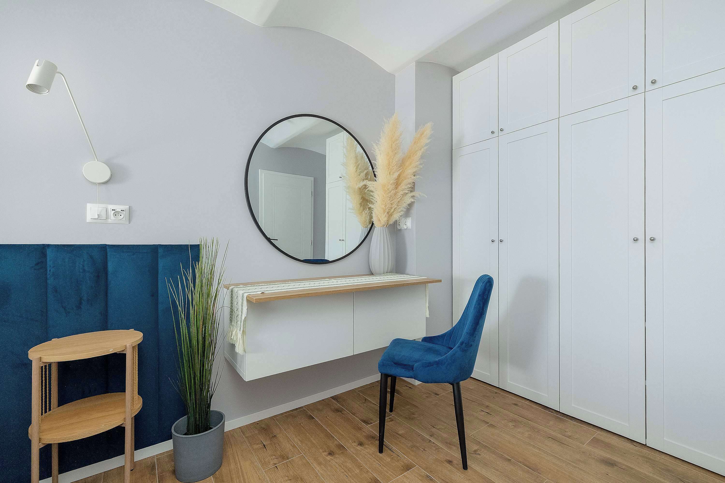 Ein Zimmer mit Holzboden, blauem Sessel, Schreibtisch aus Holz, Spiegel, weißem Kleiderschrank und einer Zimmerpflanze.