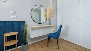 Una habitación con suelo de madera, una silla azul, un escritorio de madera, un espejo, un armario blanco y una planta en maceta.