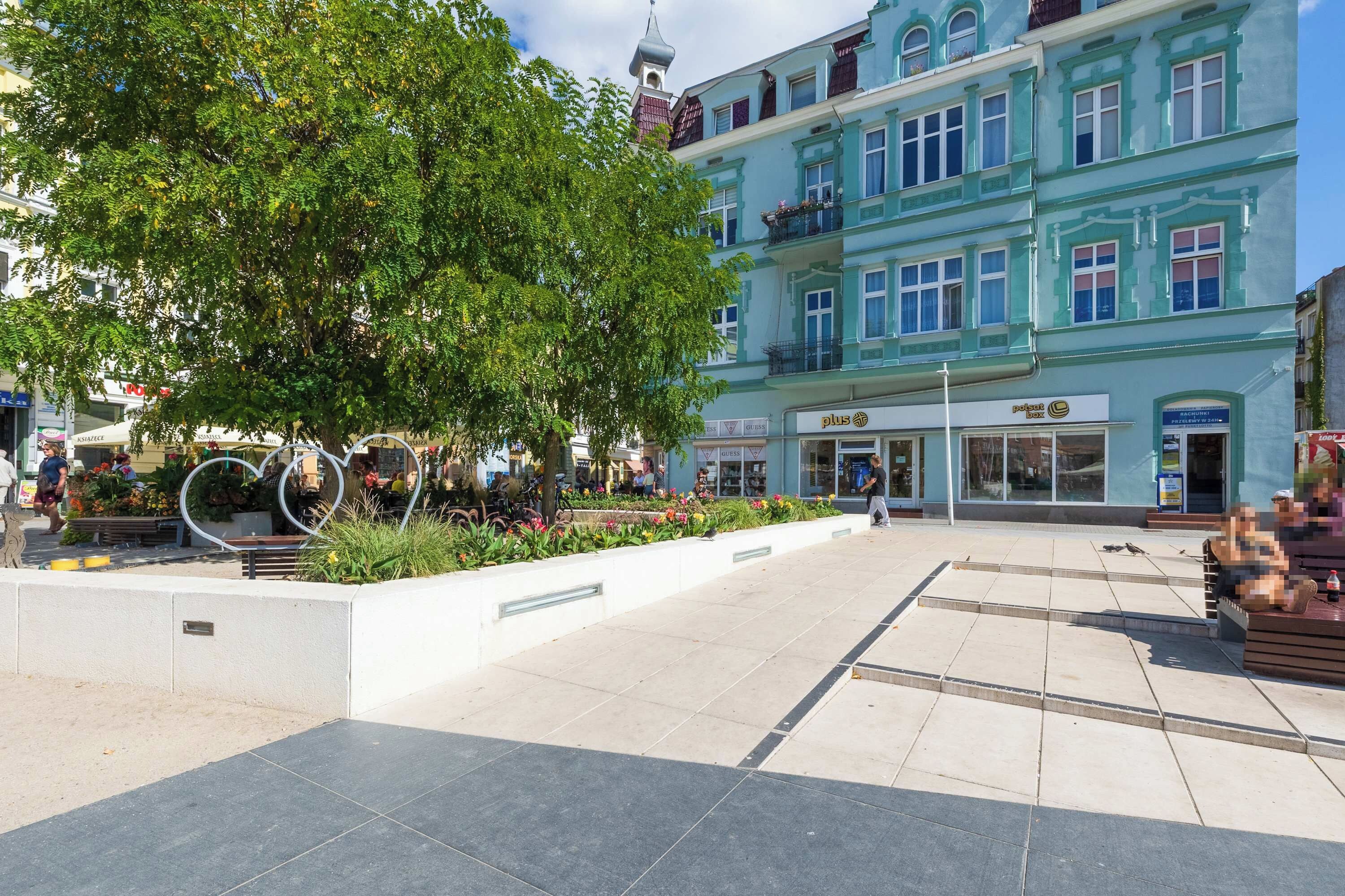 Une place publique avec un grand arbre, des bancs et un bâtiment sur lequel on peut voir un panneau avec le signe « Plus ».