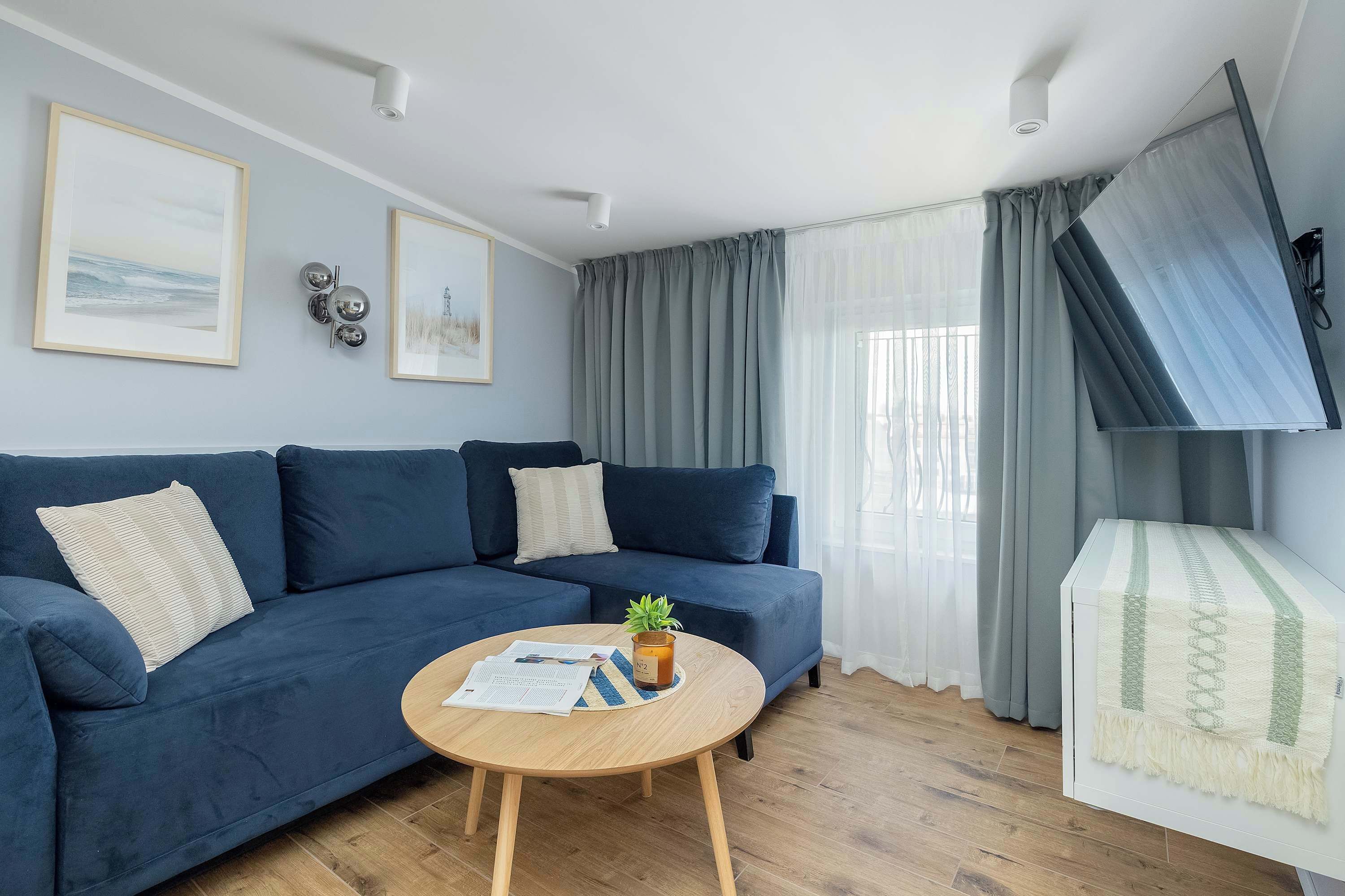 Ein Wohnzimmer mit einem blauen Sofa, einem Holztisch mit Pflanze und einem an der Wand befestigten Fernseher.