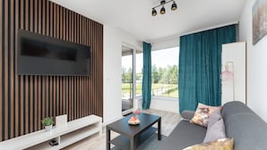 Una sala de estar moderna con un televisor de pantalla plana, un sofá con almohadones decorativos, una mesita de centro y vistas al exterior a través de grandes ventanales.
