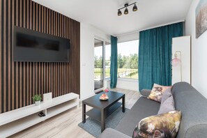 A modern living room with a flat-screen TV, a sofa with decorative pillows, a coffee table, and a view of the outdoors through large windows. - Air Conditioned Apartment with Balcony | Parking (Mielno)