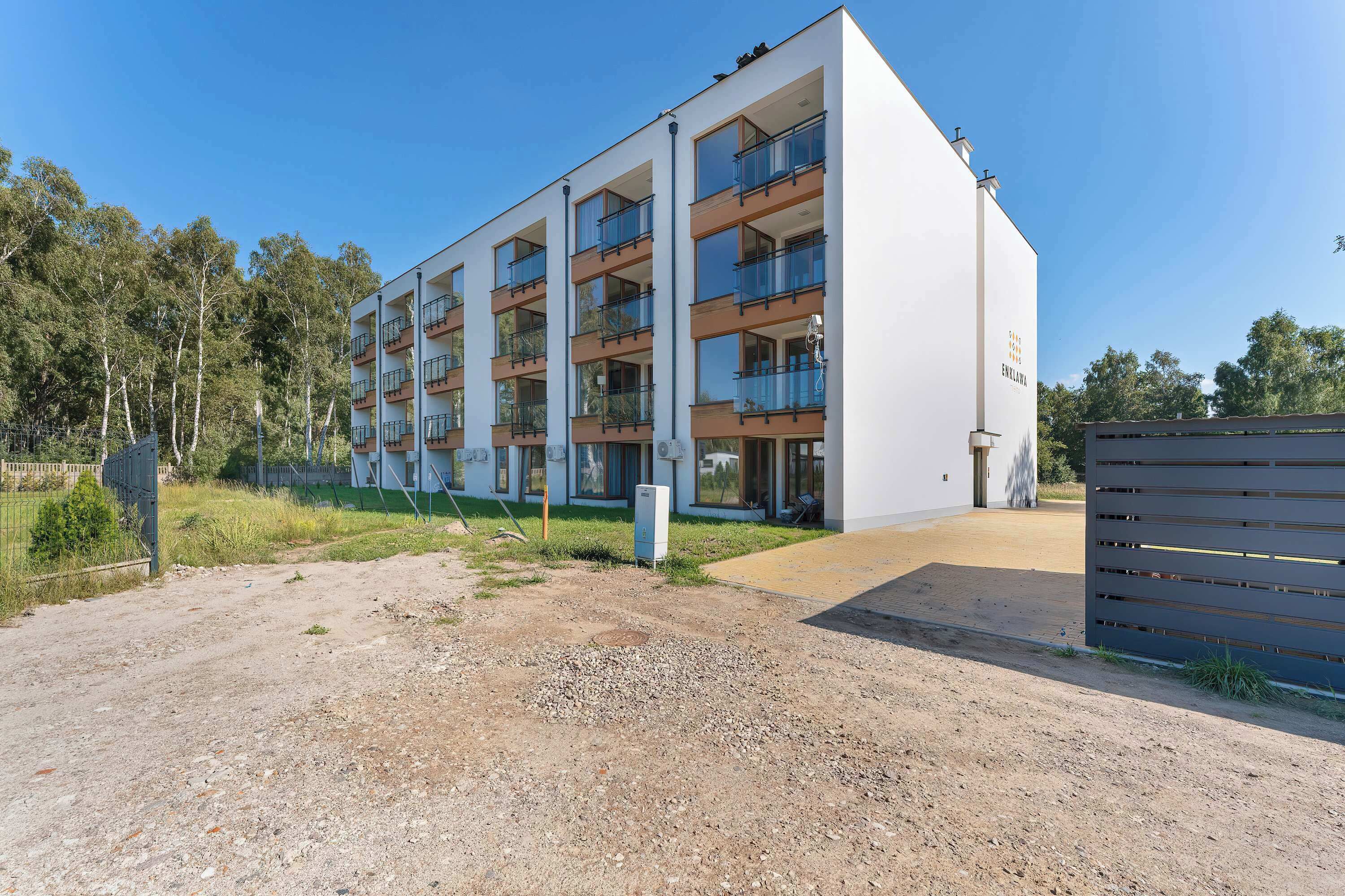 A modern multi-story residential building with balconies, a gated entrance, and a gravel driveway.