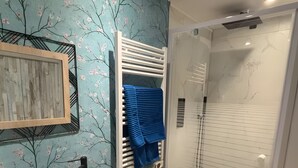 Un baño con lavamanos, un cuadro enmarcado, un perchero con una toalla azul, una ducha con puerta de vidrio y un cubo de basura.