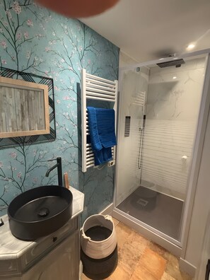 A bathroom with a sink, a framed picture, a towel rack with a blue towel, a shower with a glass door, and a trash bin.