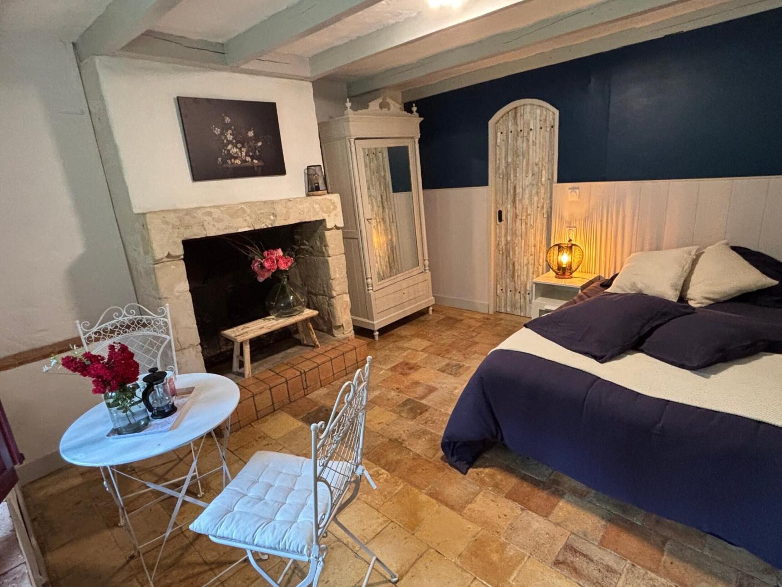 A bedroom with a bed, a fireplace, a small table with a chair, and a wardrobe.