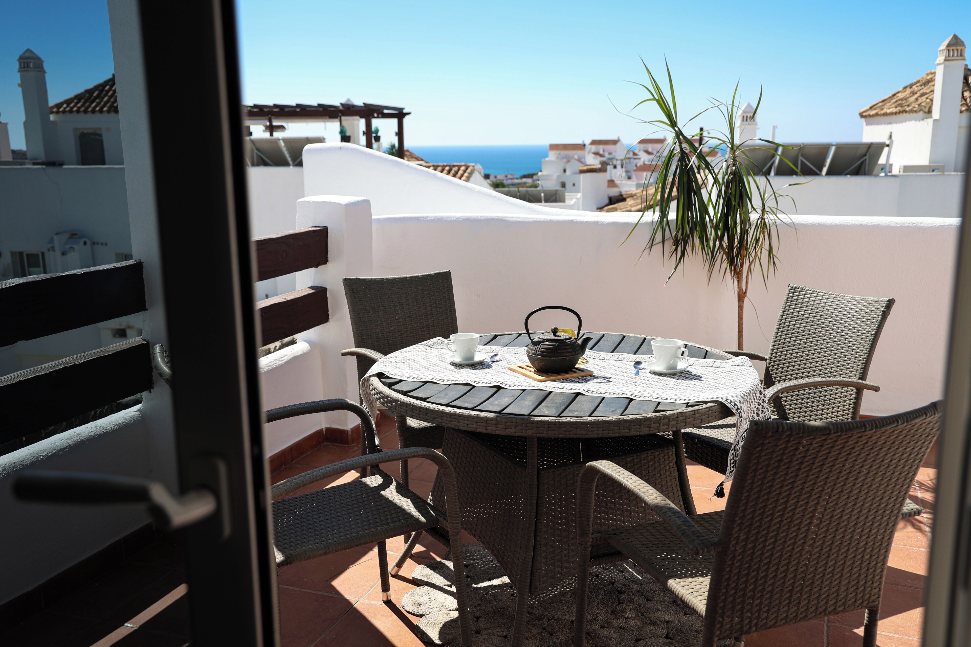 Une table ronde entourée de chaises, un pot de fleurs et une théière sur une terrasse offrant une vue sur la mer.