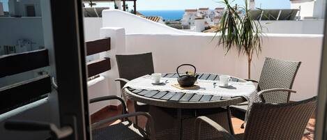 Une table ronde entourée de chaises, un pot de fleurs et une théière sur une terrasse offrant une vue sur la mer.