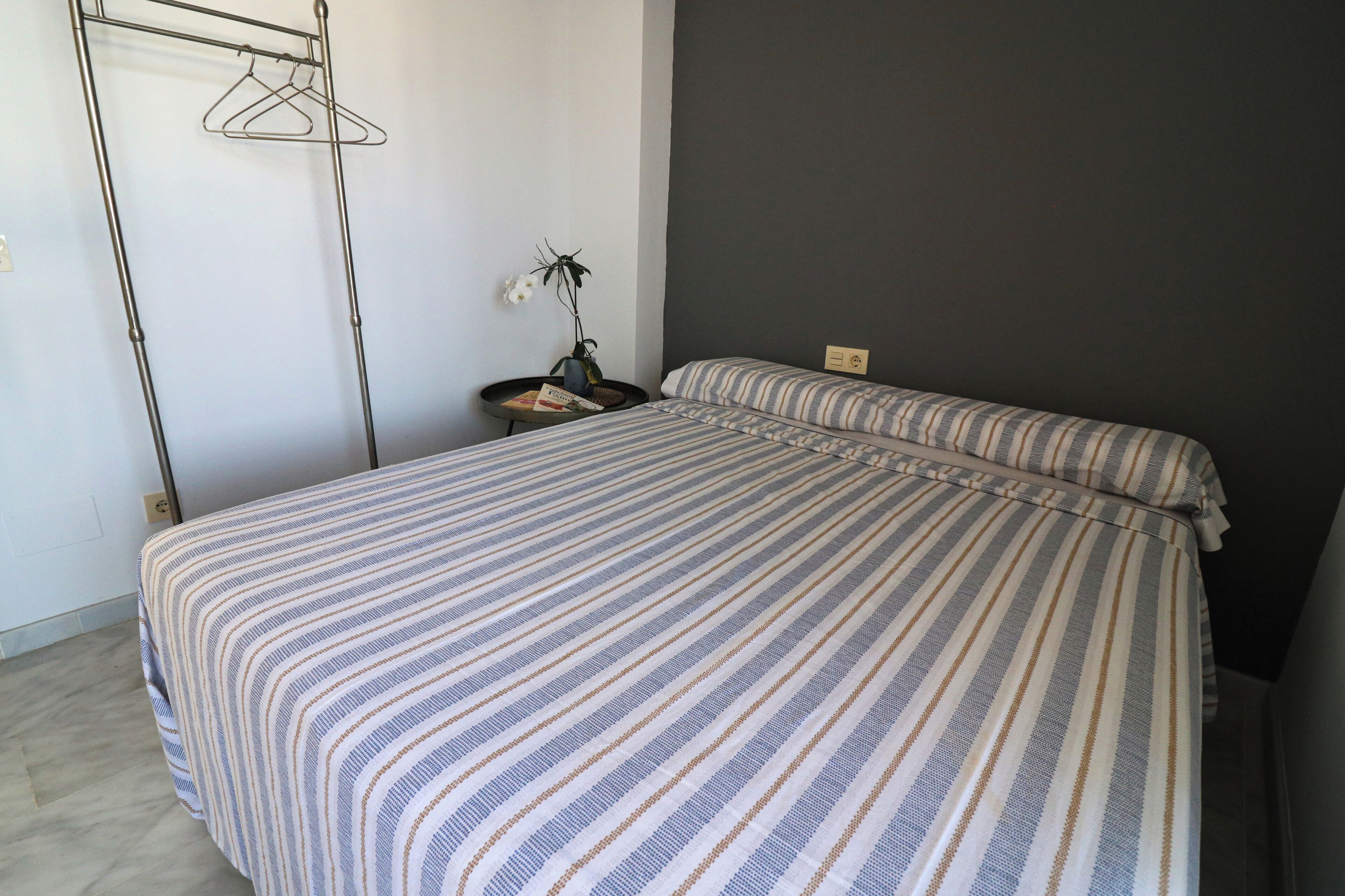 Un lit avec une literie à rayures, une petite table de chevet ornée d’un vase de fleurs et un luminaire fixé au mur.