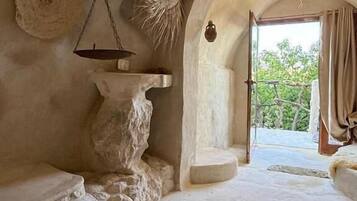 Une pièce rustique avec un évier en pierre, une balance suspendue, un miroir rond et une vue sur l’extérieur, accessible par une arche.