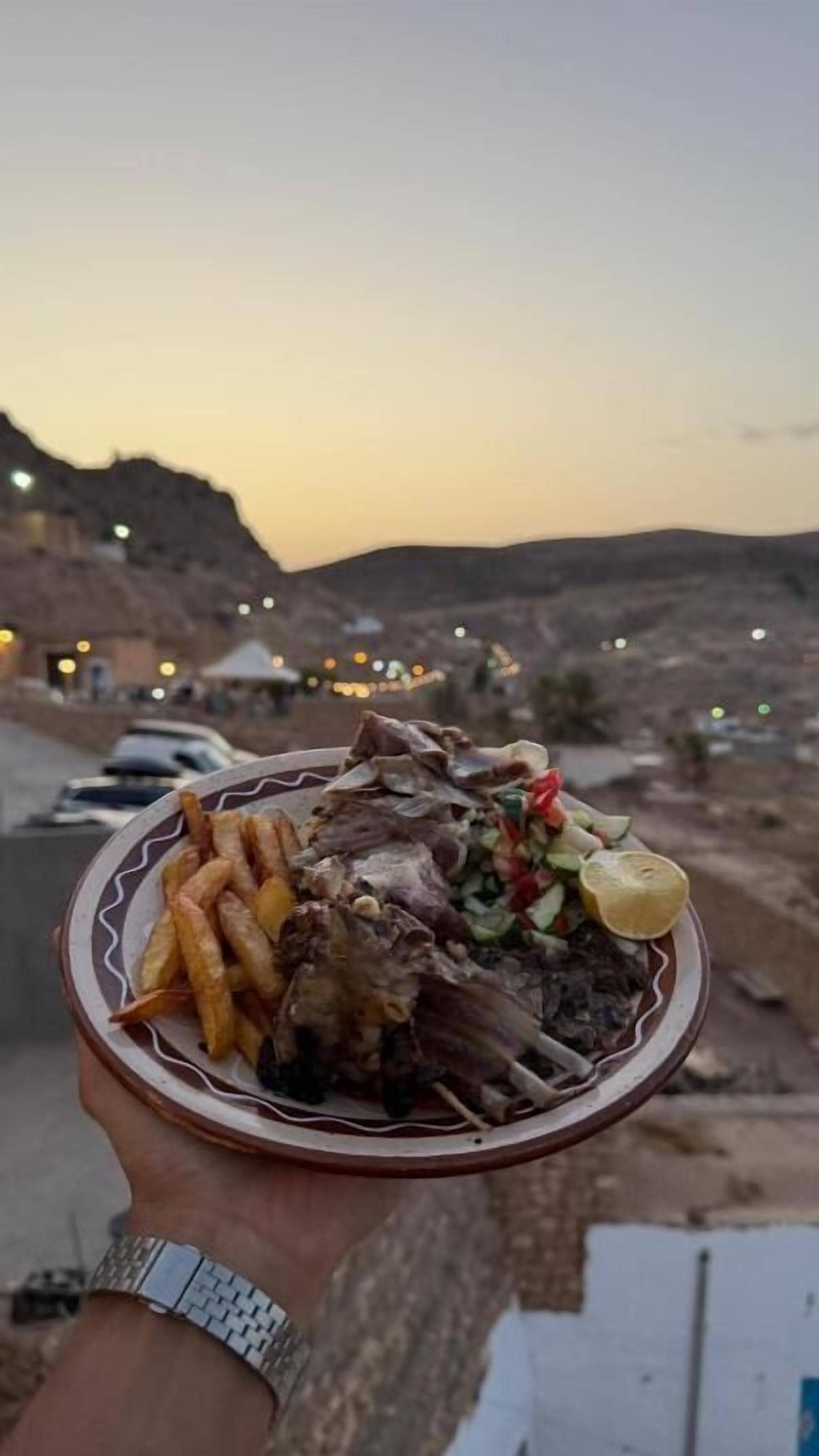 Una persona sostiene un plato con comida, un paisaje desértico y una puesta de sol al fondo.