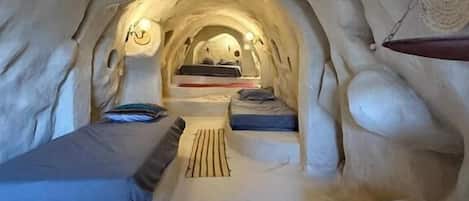 An ice hotel interior with a central arched passageway, beds, and a woven wall hanging.