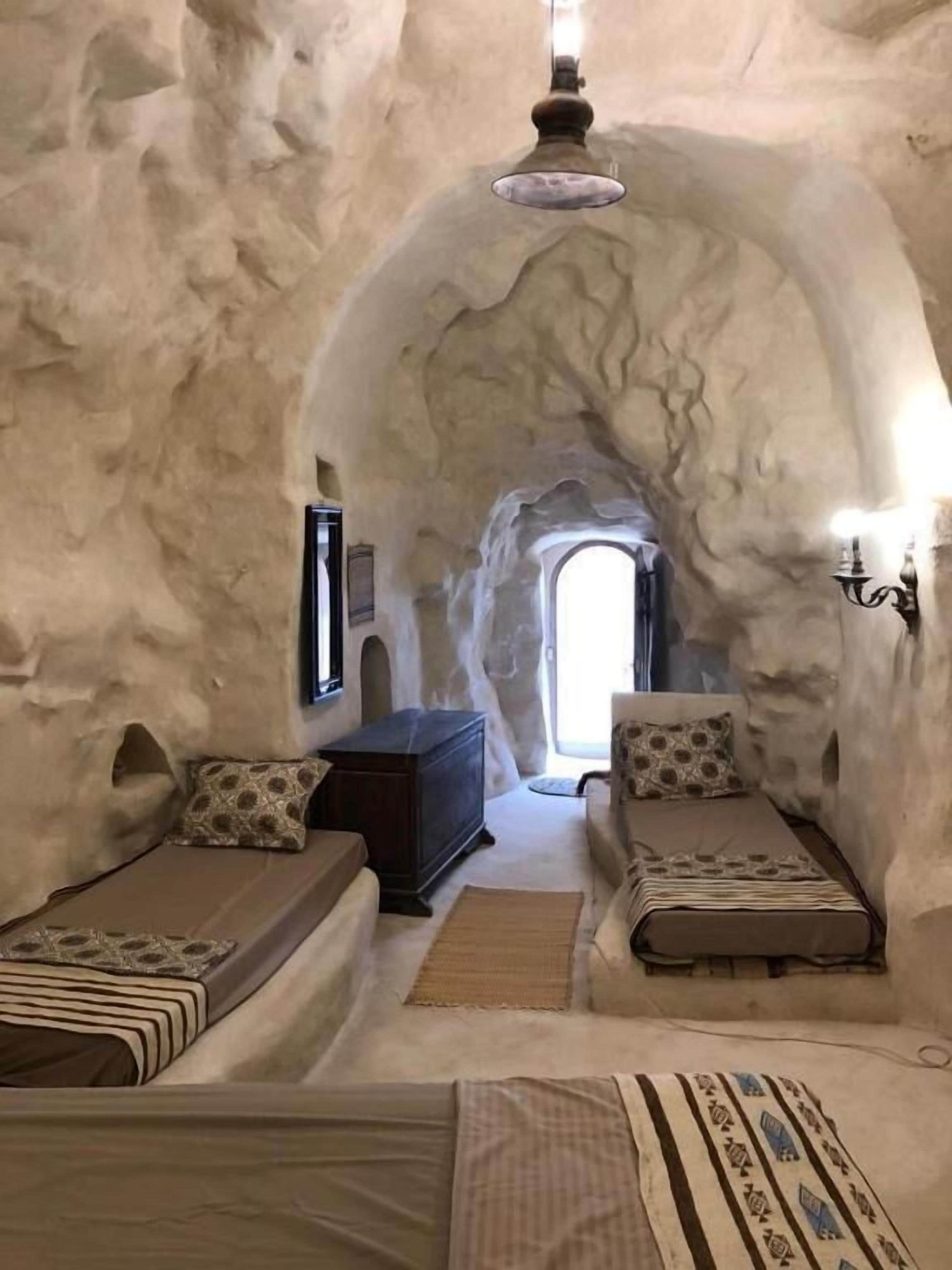 A room with a unique arched ceiling, two beds with patterned bedding, a wooden chest, and a small table.