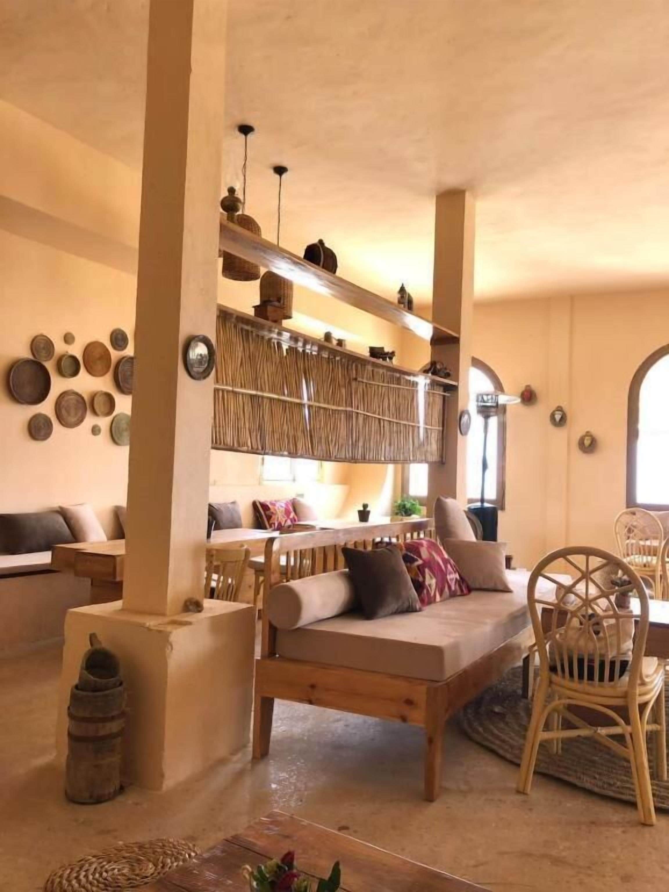 Una sala acogedora con un sofá, una silla de madera y una mesa de comedor. Se observan estanterías con objetos decorativos y un ventanal grande.