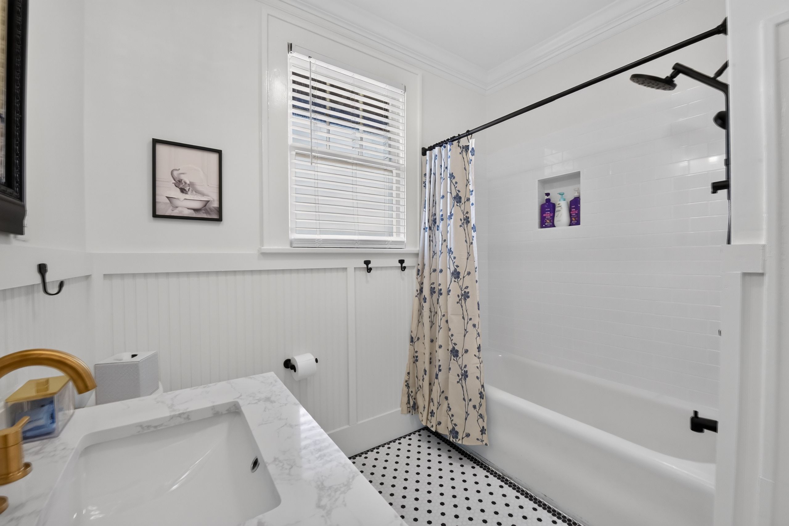 A bathroom with a bathtub, a shower, a toilet, a window with blinds, and a framed picture on the wall.