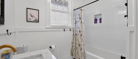 A bathroom with a bathtub, a shower, a toilet, a window with blinds, and a framed picture on the wall.