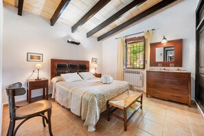 Ein Schlafzimmer mit einem Bett, Nachttischen, einem Sessel, einem Schminktisch, einem Spiegel und einem Fenster mit Vorhängen.