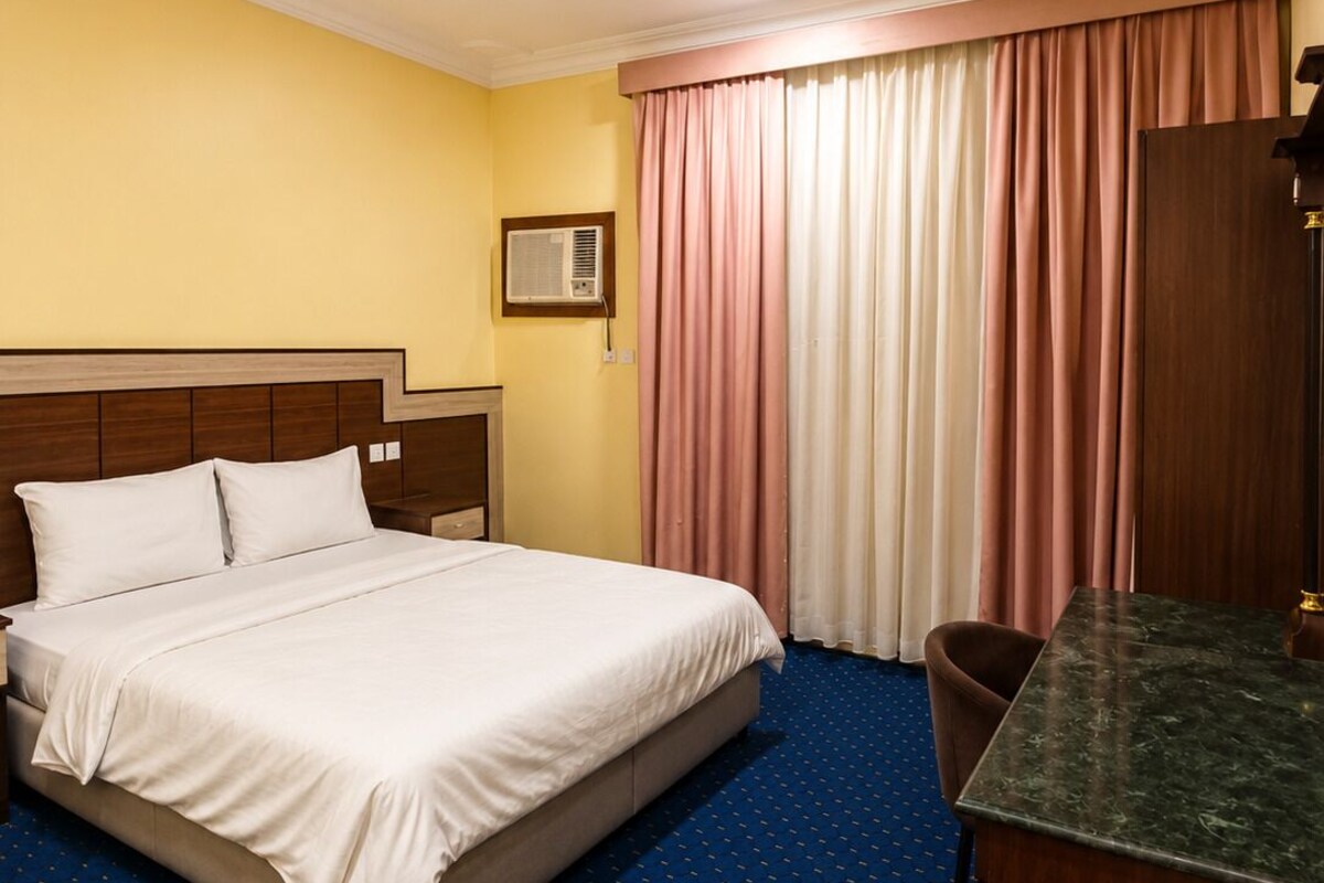 Una habitació d'hotel amb llit, escriptori, cadira, finestra amb cortines i llum al sostre.