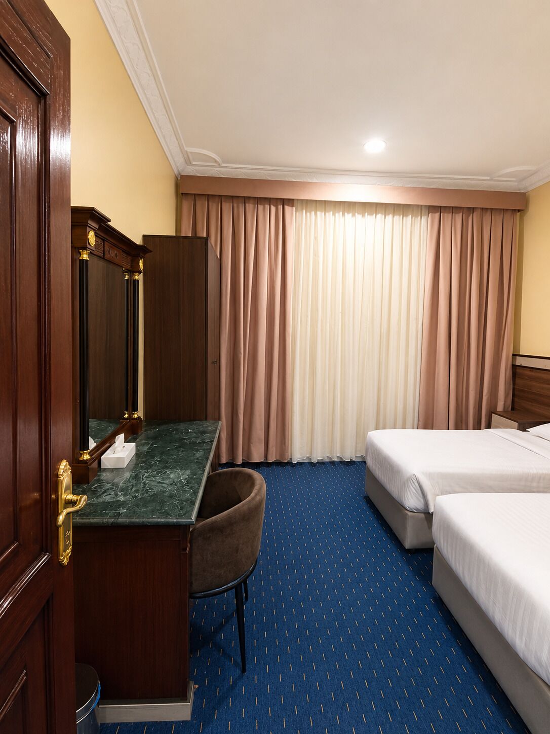 A hotel room with a marble desk, a chair, two beds, and a large window with curtains.