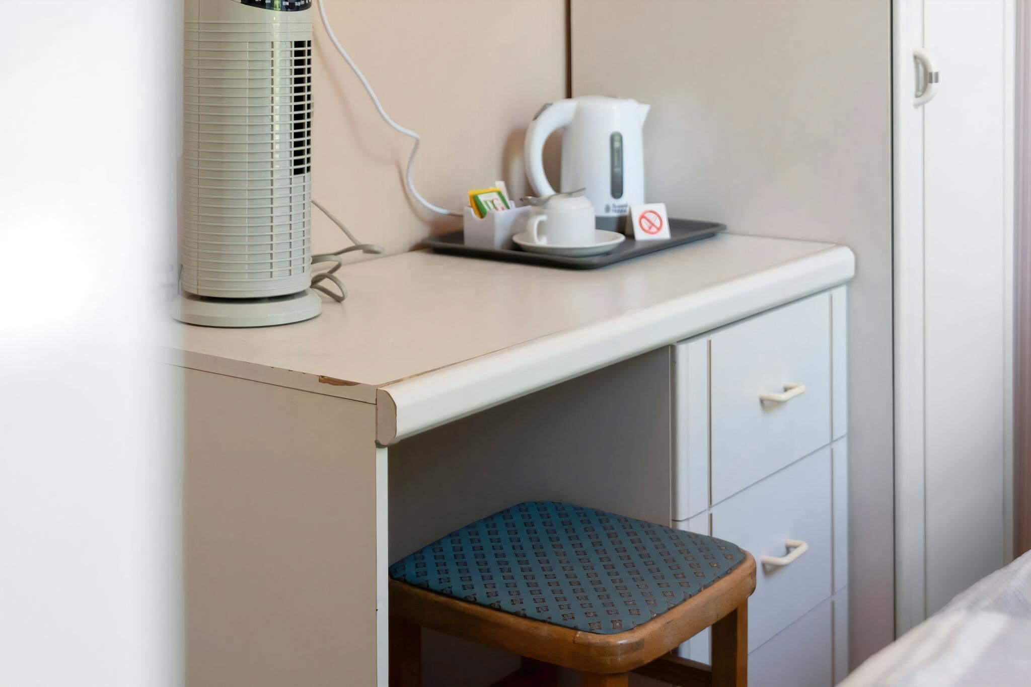 A small kitchenette with a white kettle, a cup, and a tray, a white radiator, and a wooden stool.