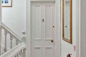 Una puerta blanca con el número 3, un espejo con marco dorado y un extintor rojo en la pared.