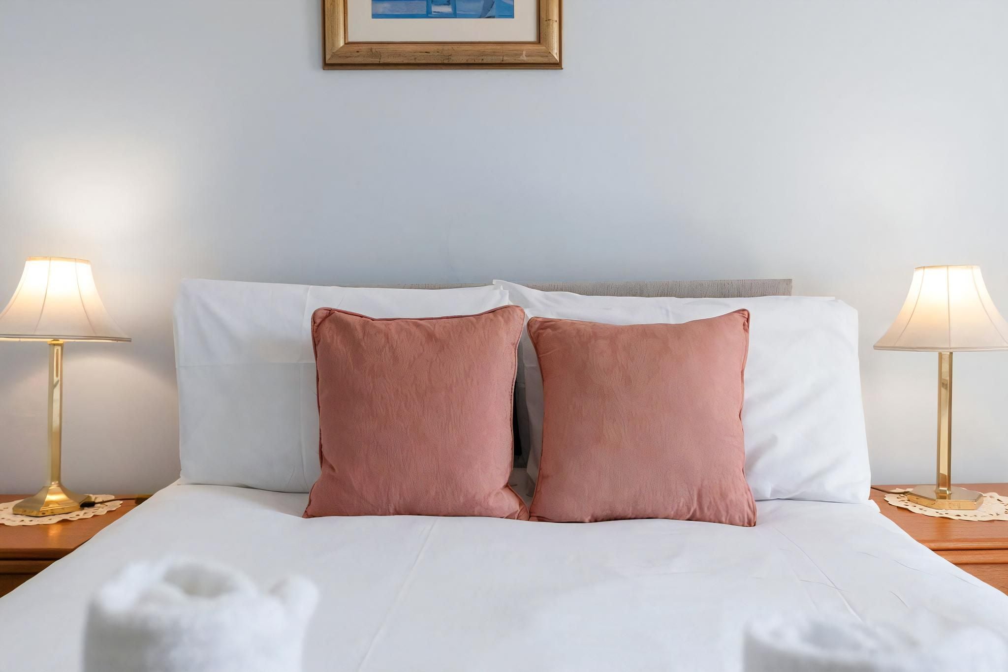 Una cama bien hecha con sábanas blancas y dos almohadas rosas, flanqueada por dos mesitas de noche con lámparas, frente a una pared lisa con un cuadro enmarcado encima.