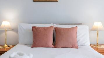 Una cama bien hecha con sábanas blancas y dos almohadas rosas, flanqueada por dos mesitas de noche con lámparas, frente a una pared lisa con un cuadro enmarcado encima.