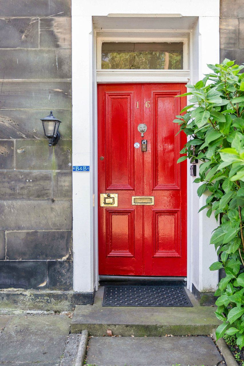 Une porte rouge avec une plaque portant un numéro, un paillasson et une petite plante à côté.