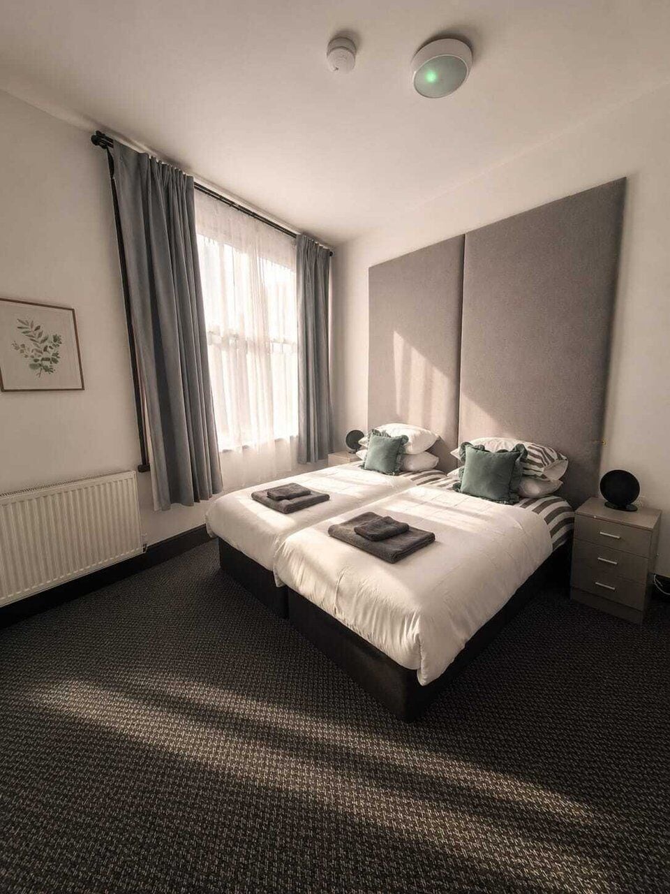 Una habitación de hotel moderna con una cama grande, una mesita de noche, un radiador y una ventana con cortinas.