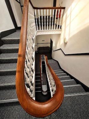 Una escalera con barandilla de madera y balaustradas blancas, escalones alfombrados y una lámpara fijada a la pared.