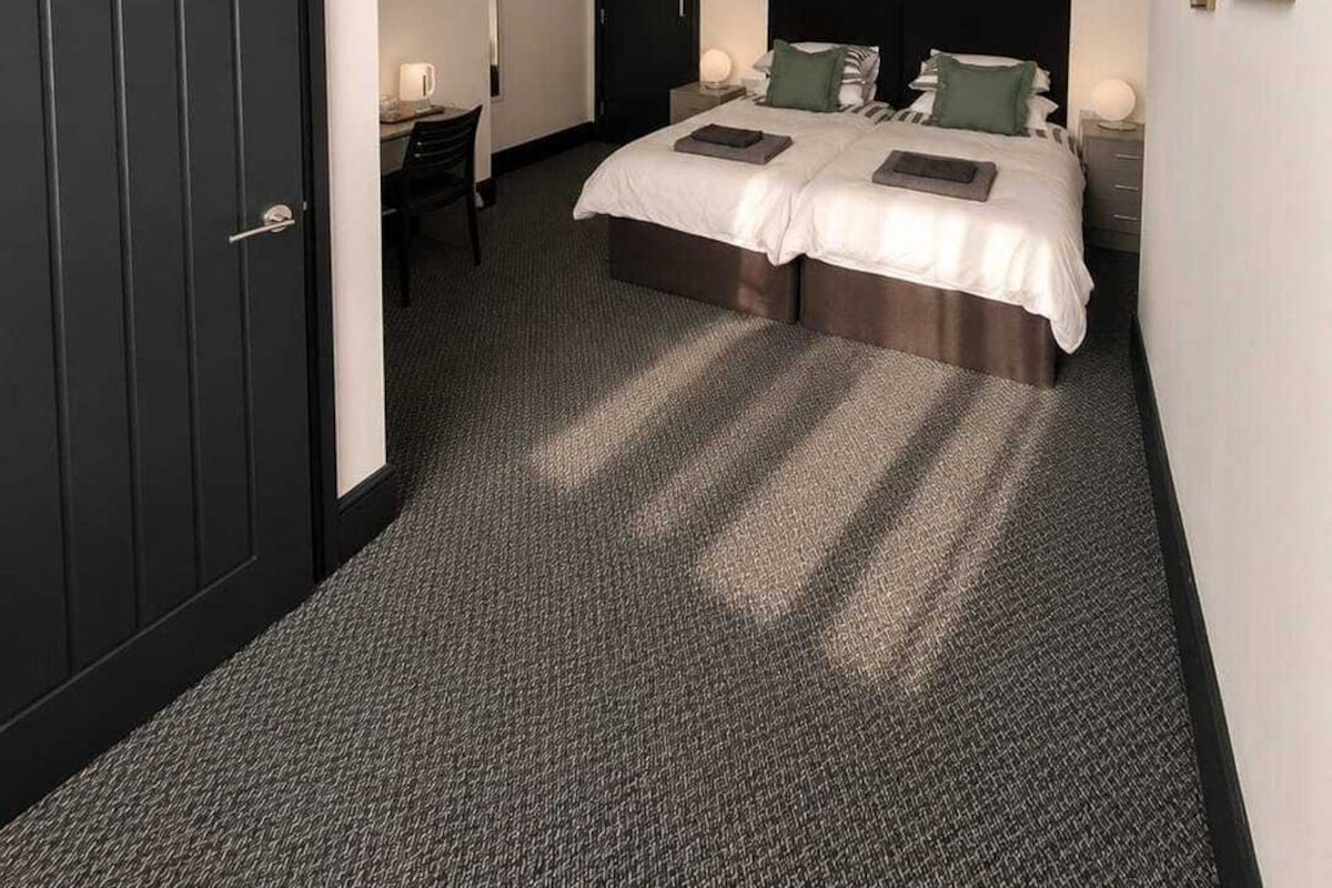 Habitación de hotel con cama doble, un escritorio con silla, perchero y una puerta oscura.