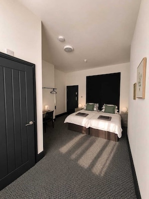 Quarto de hotel com cama de casal, uma mesa com cadeira, um cabideiro e uma porta escura.