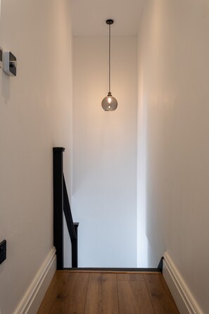 A narrow hallway with a wooden floor, a black staircase, and a hanging light fixture. - Perfect Booking 2 Bed Flat Near Cabot Circus (Bristol)