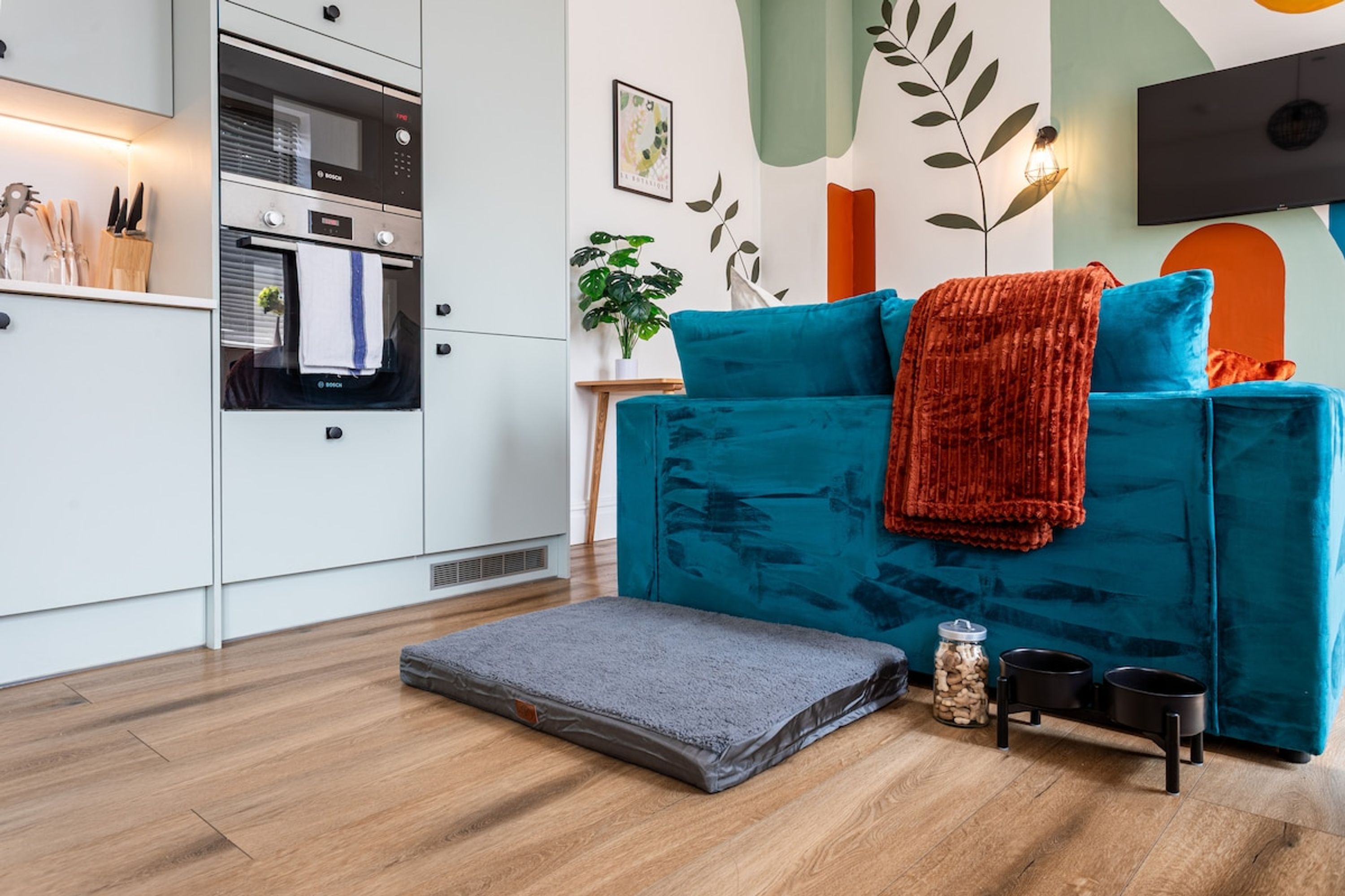 A modern kitchen with white cabinets, a Bosch oven, and a small dining area with a teal sofa, a red throw, and a grey rug.