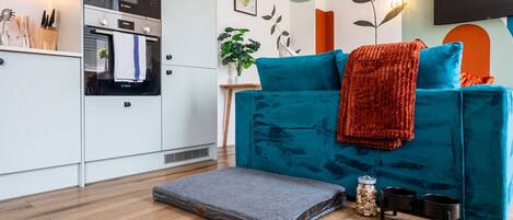 A modern kitchen with white cabinets, a Bosch oven, and a small dining area with a teal sofa, a red throw, and a grey rug.