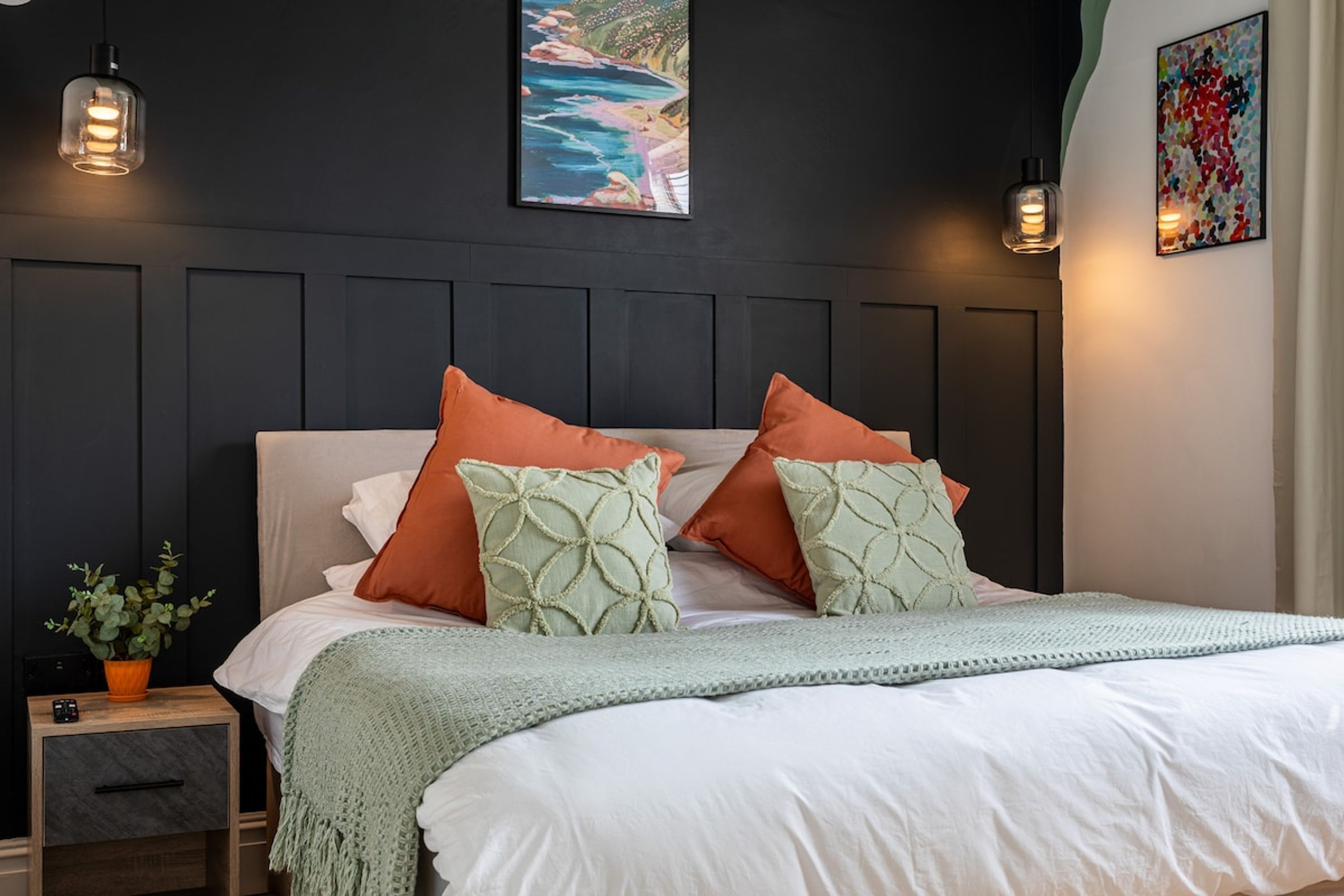 Un dormitorio con cama, almohadas, mesita de noche, una planta y decoración en la pared.