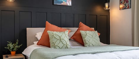 Un dormitorio con cama, almohadas, mesita de noche, una planta y decoración en la pared.